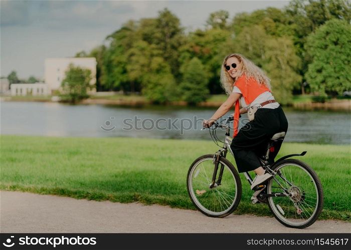 Photo of cheerful woman dressed casually, rides bicycle, looks aside, has happy expression, wears shades, poses near river, green lawn and trees, some buildings in background. People and rest