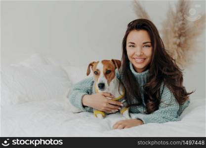 Photo of cheerful woman embraces dog with love, spend free time together, expresses tender feeling and emotions, falls in love with pet, lie on comfortable bed. Positive emotions, animals and care