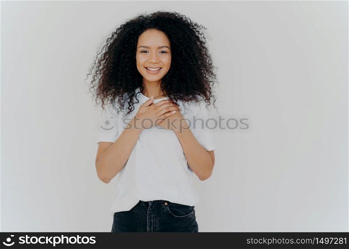 Photo of cheerful woman with Afro hair, keeps hands on chest, expresses gratitude, smiles gently, wears white t shirt and jeans, impressed to get pleasant compliment, isolated over white background