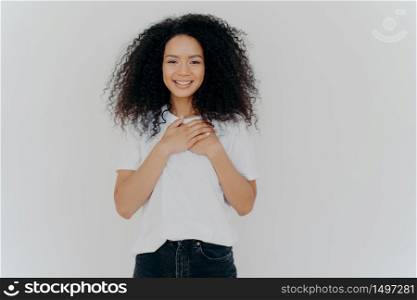 Photo of cheerful woman with Afro hair, keeps hands on chest, expresses gratitude, smiles gently, wears white t shirt and jeans, impressed to get pleasant compliment, isolated over white background