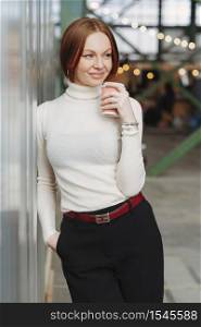 Photo of cheerful young woman wears elegant clothes, dressed in white jumper and black trousers, drinks disposable cup of coffee, dreams about something pleasant, keeps gaze aside. Rest concept