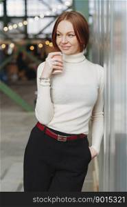 Photo of cheerful young woman wears elegant clothes, dressed in white jumper and black trousers, drinks disposable cup of coffee, dreams about something pleasant, keeps gaze aside. Rest concept