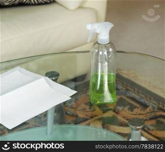 Photo of cleaning solution, in spray bottle, on dirty glass table with paper towels and sofa in background