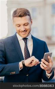 Photo of clever male lawyer waits for client outdoor, looks at wristwatch, holds smart phone as waits for important call, wears formal suit, has pleasant smile on face. People, time, meeting concept