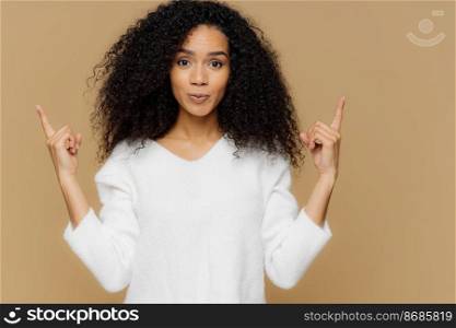Photo of confident woman has bushy Afro hairstyle, points both index fingers above, dressed in white jumper, stands against brown background, advertises some item, shows direction. Come upstairs