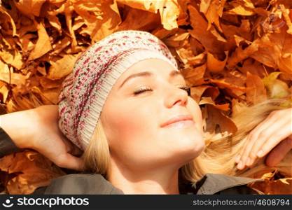 Photo of cute blond girl dreaming on the ground covered orange autumnal leaves in the garden, pretty woman relaxed in autumn park, attractive female enjoying warm fall sunlight, joy concept