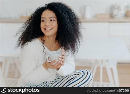 Photo of cute cheerful woman with bushy curly hair looks away with smile, holds mug of coffee, wears casual clothing, enjoys cozy calm morning, savors taste. Afro lay in kitchen during day off