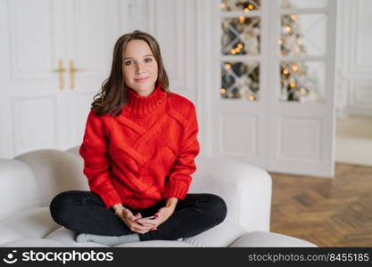 Photo of cute European woman has dark hair, dressed in domestic clothes, spends winter weekend at home, sits in lotus pose on comfortable sofa, uses moden cell phone for online communication