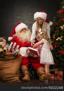 Photo of cute girl and her mother and Santa Claus holding giftbox and opening it at home near Christmas tree