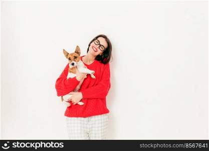 Photo of delighted Caucasian female carries jack russell terrier dog, wears spectacles and red sweater, enjoys spare time with favourite pet, pose against white background. People and animals concept