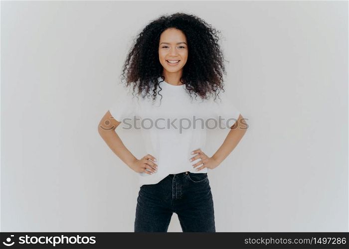 Photo of delighted curly woman keeps both hands on waist, smiles gently, has slim figure, wears white t shirt and black jeans, being in good mood, stands self assured against white background