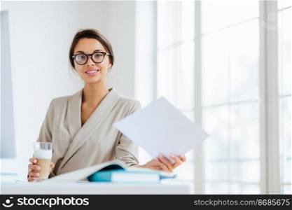 Photo of delighted dark haired European woman in transparent glasses, works with documents, reads information data, dressed in elegant clothing, drinks milkshake sits at desktop in office makes report