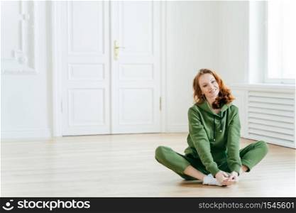 Photo of delighted redhead female sits on lotus pose, wears tracksuit, being in good body shape, has curly ginger hair, poses on floor, copy space for your promotional content. Sport concept