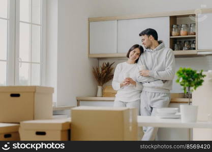 Photo of delighted relaxed husband and wife pose near modern kitchen furniture, have glad expressions, drink takeaway coffee, surrounded with cardboard boxes during relocation day. Mortgage.