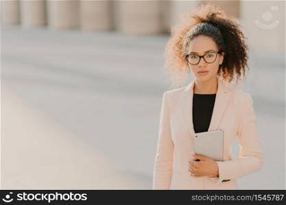 Photo of elegant African American businesswoman uses tablet computer outdoor, wears formal white jacket, transparent glasses with black frames, curly hair combed in pony tail, blank space on left