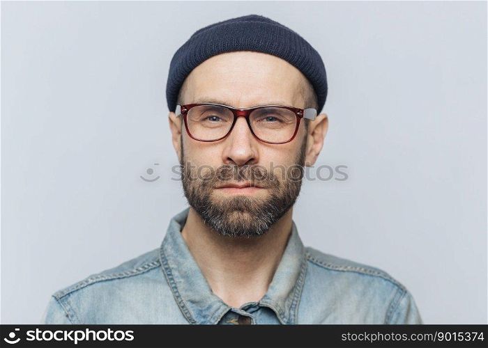 Photo of∫elli≥nt confident stylish man with dark thick beard andμstache, looks seriously∫o camera, poses against grey studio background, wears spectac≤s and hat. Facial expressions concept