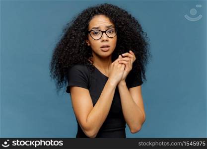 Photo of embarrassed Afro American female keeps hands together, looks nervously at camera, has some problems, wears casual black t shirt, stands against blue background. Negative feelings concept