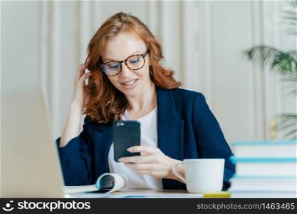 Photo of female freelancer uses smart phone for online communication, has red curly hair, wears optical glasses and formal costume, has coffee break after working on laptop computer, uses free wifi