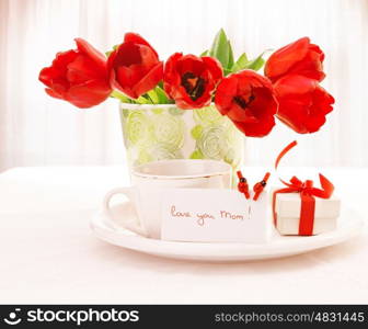 Photo of fresh red tulip flowers in beautiful vase on table, cup of tea, little white gift box with ribbon, festive postcard with ladybird decorations, home interior, happy mothers day, love concept