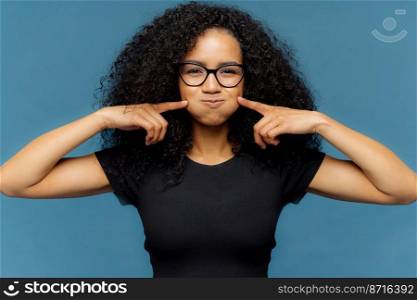 Photo of funny merry female blows cheeks, touches with both index fingers, wears black casual t shirt, optical eyewear, stands over blue background, makes grimace and foolishes indoor alone.