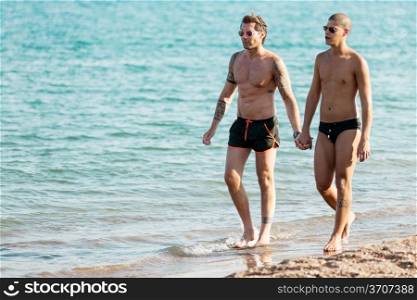Photo of gay couple at the beach walking through the water while holding hand &#xA;