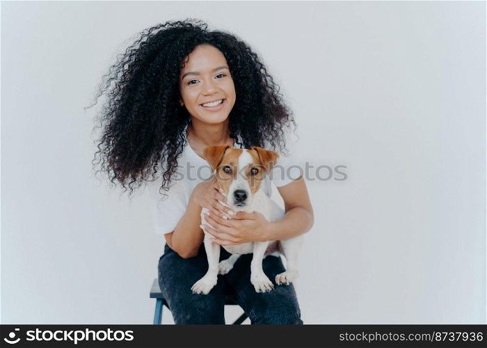 Photo of glad dark skinned female being forever together with pedigree puppy, enjoys leisure at home, sits on chair, isolated over white background. Human and animal connection. Trust and friendship
