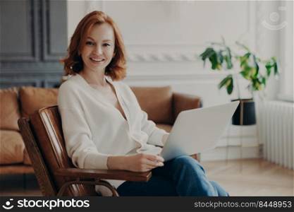 Photo of glad European woman freelancer satisfied with remote job, works freelance on laptop computer, poses on cozy room with modern furniture, works online with documents, dressed casually