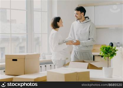 Photo of glad husband and wife carry plates, pose in kitchen near big window, unpack personal belongings from cardboard boxes, dressed in casual outfit, smile pleasantly during talk with each other