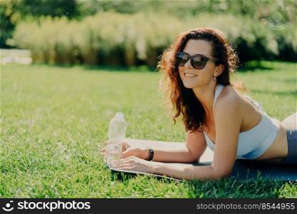 Photo of glad relaxed brunette woman poses on fitness mat wears trendy sunglasses and sportswear drinks water as feels thirsty after sport exercises lying on green grass enjoys good sunny day.