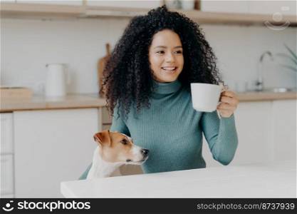 Photo of glad young woman has Afro hairstyle, holds white mug of hot drink, looks with toothy smile somewhere into distance, sits at kitchen table, spends time with favourite pet, cozy atmosphere