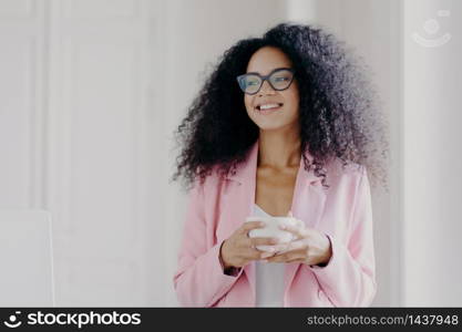 Photo of good looking African American prosperous busineswoman waits for partner in office, drinks coffee discusses future plans with colleague looks away with cheerful expression wears formal clothes