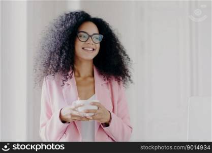 Photo of good looking African American prosperous busineswoman waits for partner in office, drinks coffee discusses future plans with colleague looks away with cheerful expression wears formal clothes