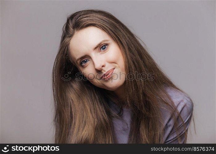 Photo of good looking blue eyed female model with dark long hair and blue appealing eyes, poses against grey background, looks with satisfied expression directly at camera. Beauty and people