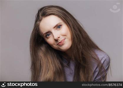 Photo of good looking blue eyed female model with dark long hair and blue appealing eyes, poses against grey background, looks with satisfied expression directly at camera. Beauty and people