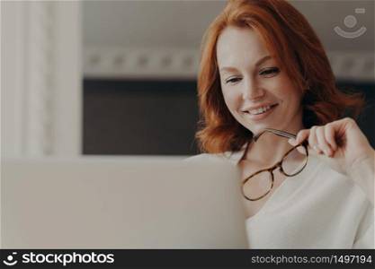 Photo of good looking cheerful redhead woman reads information on website in internet, works on laptop computer, holds transparent glasses, works on freelance project from home, shares content online
