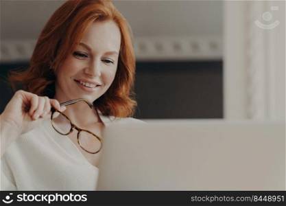 Photo of good looking cheerful redhead woman reads information on website in internet, works on laptop computer, holds transparent glasses, works on freelance project from home, shares content online
