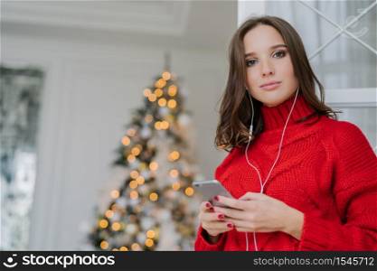 Photo of good looking female with dark hair, dressed in red knitted sweater, surfes social networks on mobile phone, listens music with earphones, admires decorated Christams tree. Leisure concept