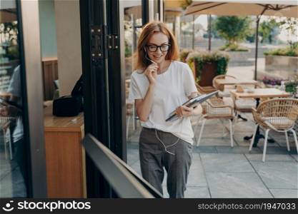 Photo of good looking ginger European woman talks via video chat app uses modern gadgets dressed in casual outfit poses against outdoor cafe background makes online call during walk in city.. Ginger European woman talks via video chat app uses modern gadgets poses outdoor