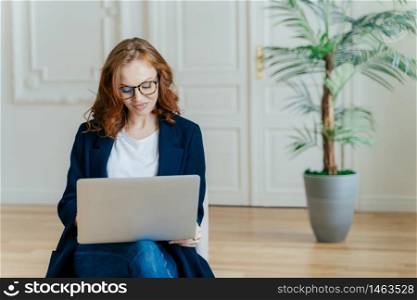 Photo of good looking ginger young woman in spectacles watches tutorial video online, poses in comfortable couch in modern apartment, dressed in elegant outfit, enjoys free internet connection