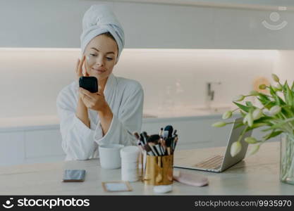 Photo of good looking woman applying face cream on face looks at herself in mirror cares about complexion wears bath towel and robe sits at table against kitchen interior. Beauty time concept