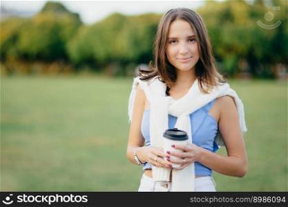 Photo of good looking woman with dark bobbed hairstyle, holds paper cup of coffee, has free time, spends weekend on nature, poses agaist green nature background with copy space for your text