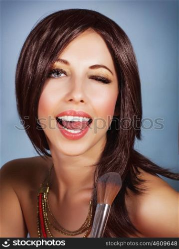Photo of gorgeous female with perfect makeup isolated on blue background, sexy brunette woman holding in hand brush for blush, playful young lady with open mouth and closed one eyes, beauty concept&#xA;
