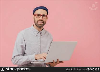 Photo of guy internet user browses website on laptop computer, connected to wireless internet, holds electronic gadget, wears spectacles, formal shirt, isolated over pink background with free space