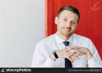Photo of handsome intelligent man with gentle smile, keeps hands together, wears formal white shirt, poses against red and white background. Male business owner with confident expression. Career