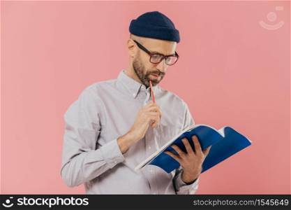 Photo of handsome male author has dark bristle, holds pencil and book, underlines necessary information, isolated over pink studio wall, has attentive gaze in notepad. Man notes something indoor