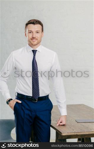Photo of handsome male office worker dressed in formal clothes, keeps one hand in pocket, other on table, looks confidently at camera, poses in office. People, business and lifestyle concept