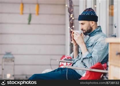 Photo of handsome male remembers something in life while drinks tea or coffee, has thoughtfull look down, recreats at terrace, wears fashionable hat and jean jacket. Leisure and people concept