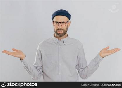 Photo of handsome man with uncertain expression, spreads hands, wears glasses and headgear, isolated over white backgrounnd. Unshaven young Caucasian male shruggs shoulders in bewilderment indoor