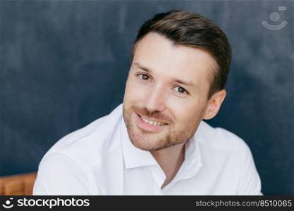 Photo of handsome unshaven male with bristle dressed in elegant white shirt, has charming smile, happy to meet with old friend, isolated over dark wall. People, emotions and lifestyle concept