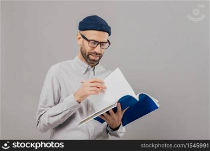 Photo of handsome unshaven young guy holds notebook, turns over page, writes records and planning for next week, wears optical glasses and white formal shirt, models over grey background alone